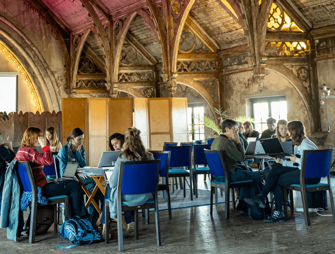 Een groep mensen zit aan tafels met laptops in een ruimte met zichtbare houten dakconstructie