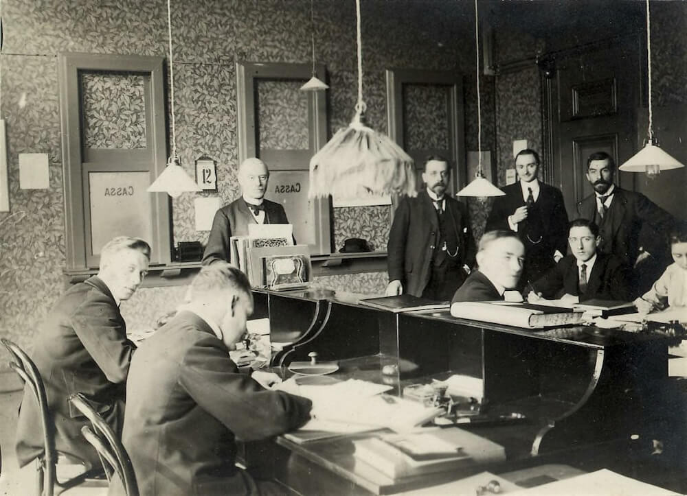 Zwartwitfoto van een kantoor in 1919 waar allerlei mannen in pakken zitten of staan