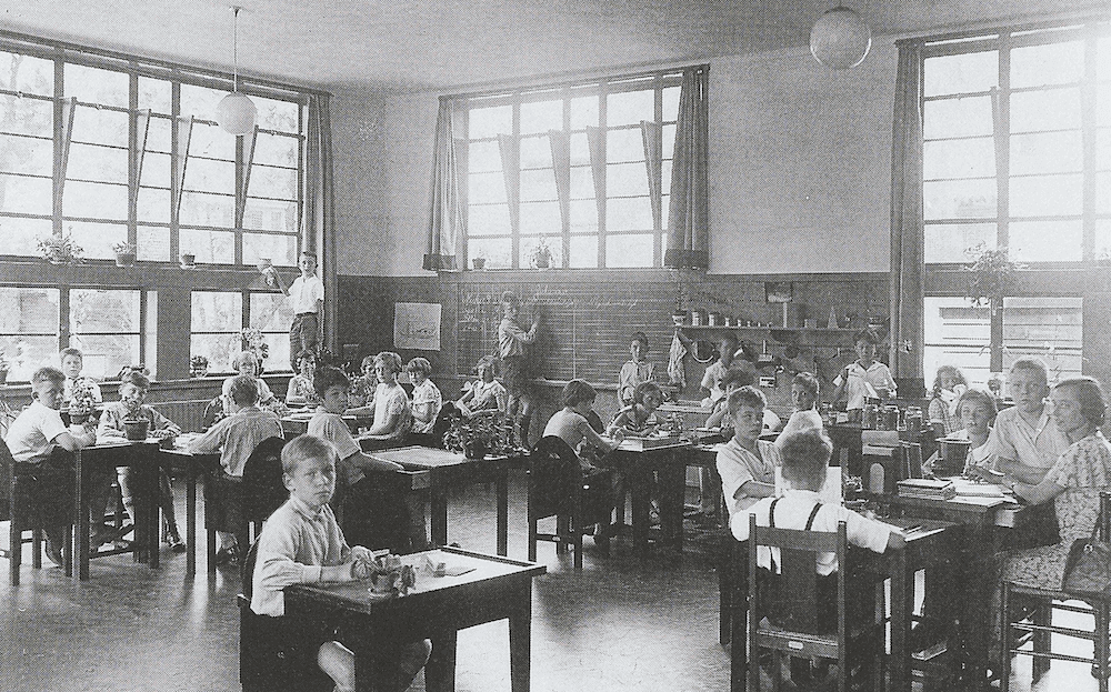 Zwartwitfoto van kinderen die verspreid aan tafels in een klaslokaal zitten.