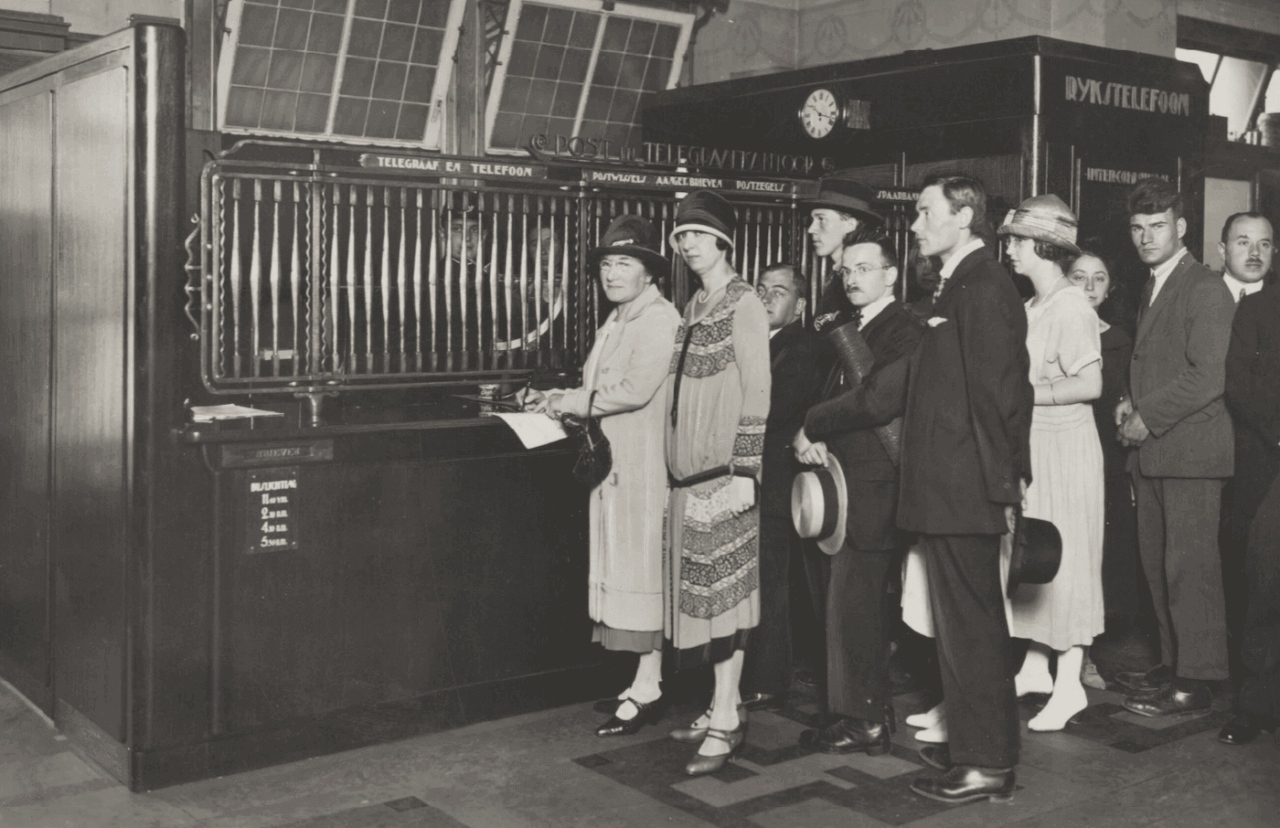 Zwartwitfoto van klanten die in een rij staan te wachten voor het loket van het pas geopende postkantoor in warenhuis de Bijenkorf, Amsterdam, 1925