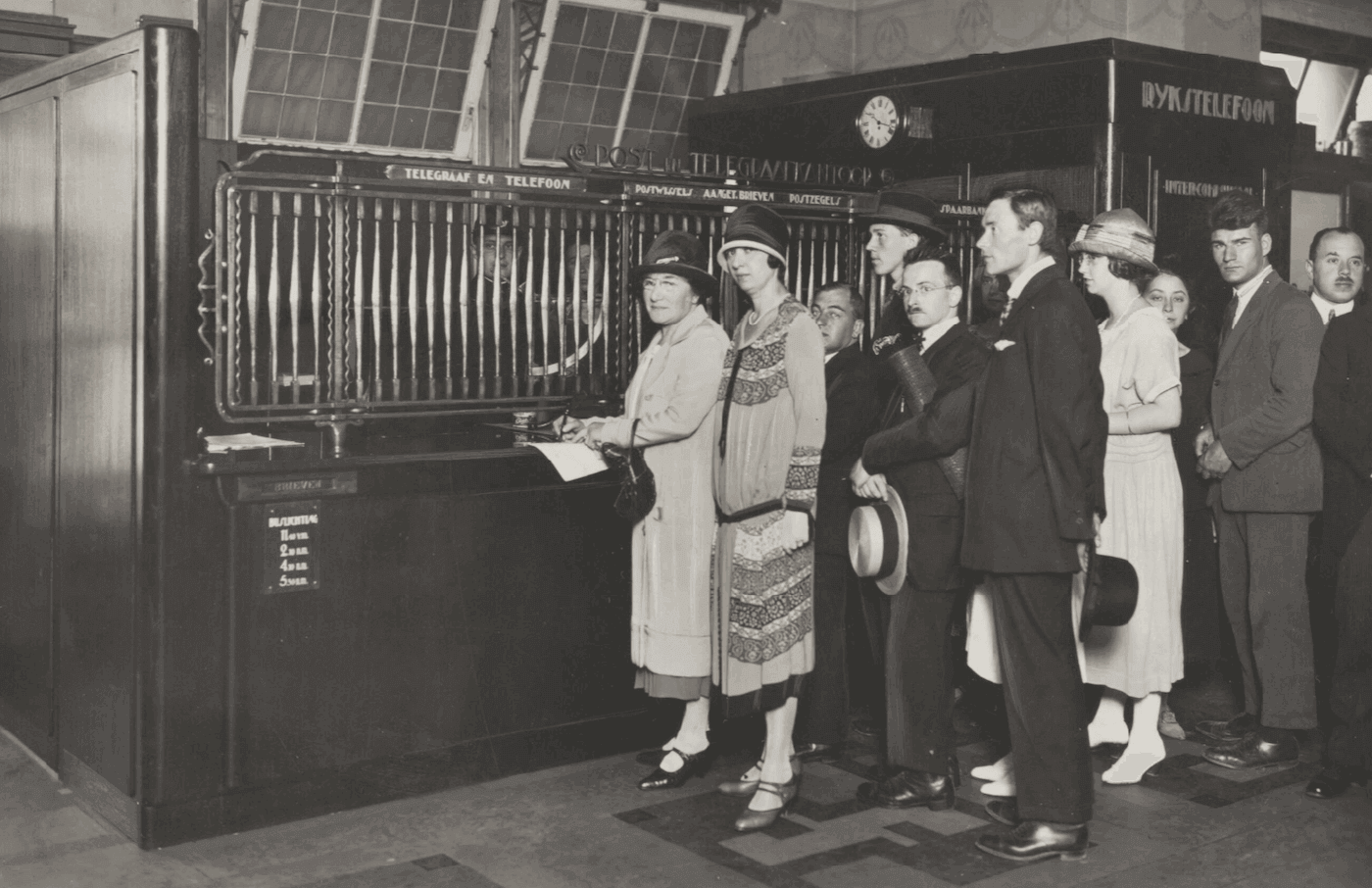 Zwartwitfoto van klanten die in een rij staan te wachten voor het loket van het pas geopende postkantoor in warenhuis de Bijenkorf, Amsterdam, 1925
