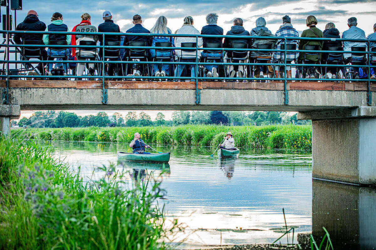 Muziektheatervoorstelling in de openlucht: twee spelers in een bootje, publiek kijkt vanaf een brug toe