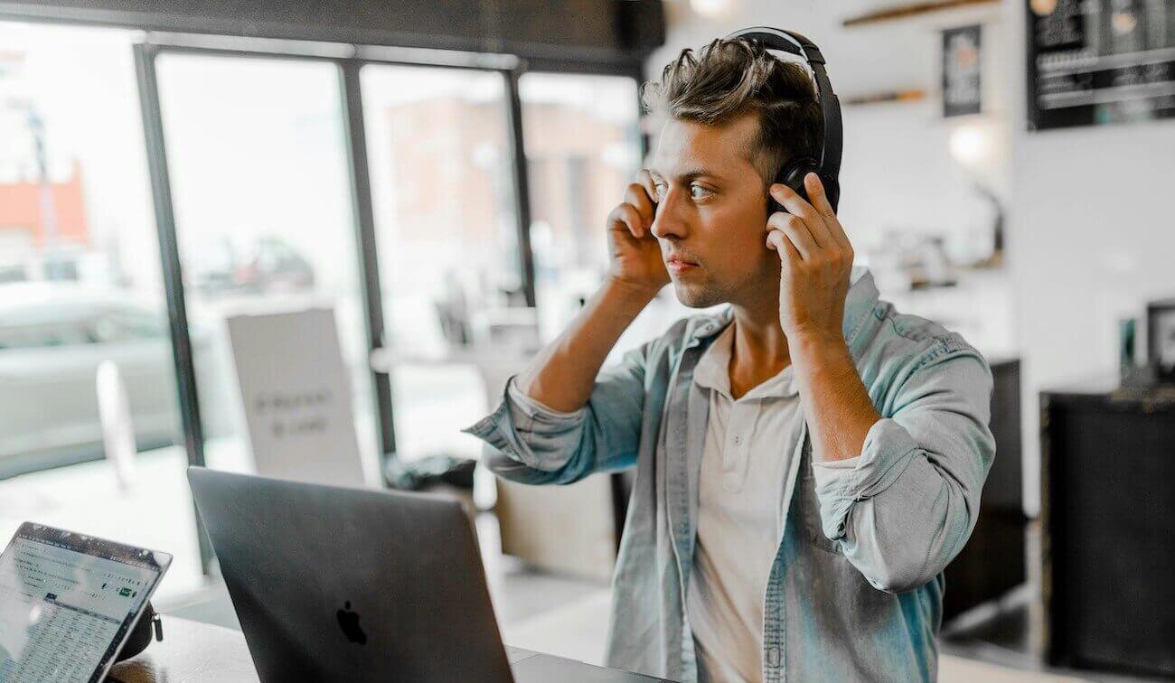 Foto van een jongeman achter een laptop in een moderne kantoorruimte die een koptelefoon opzet en naar buiten kijkt