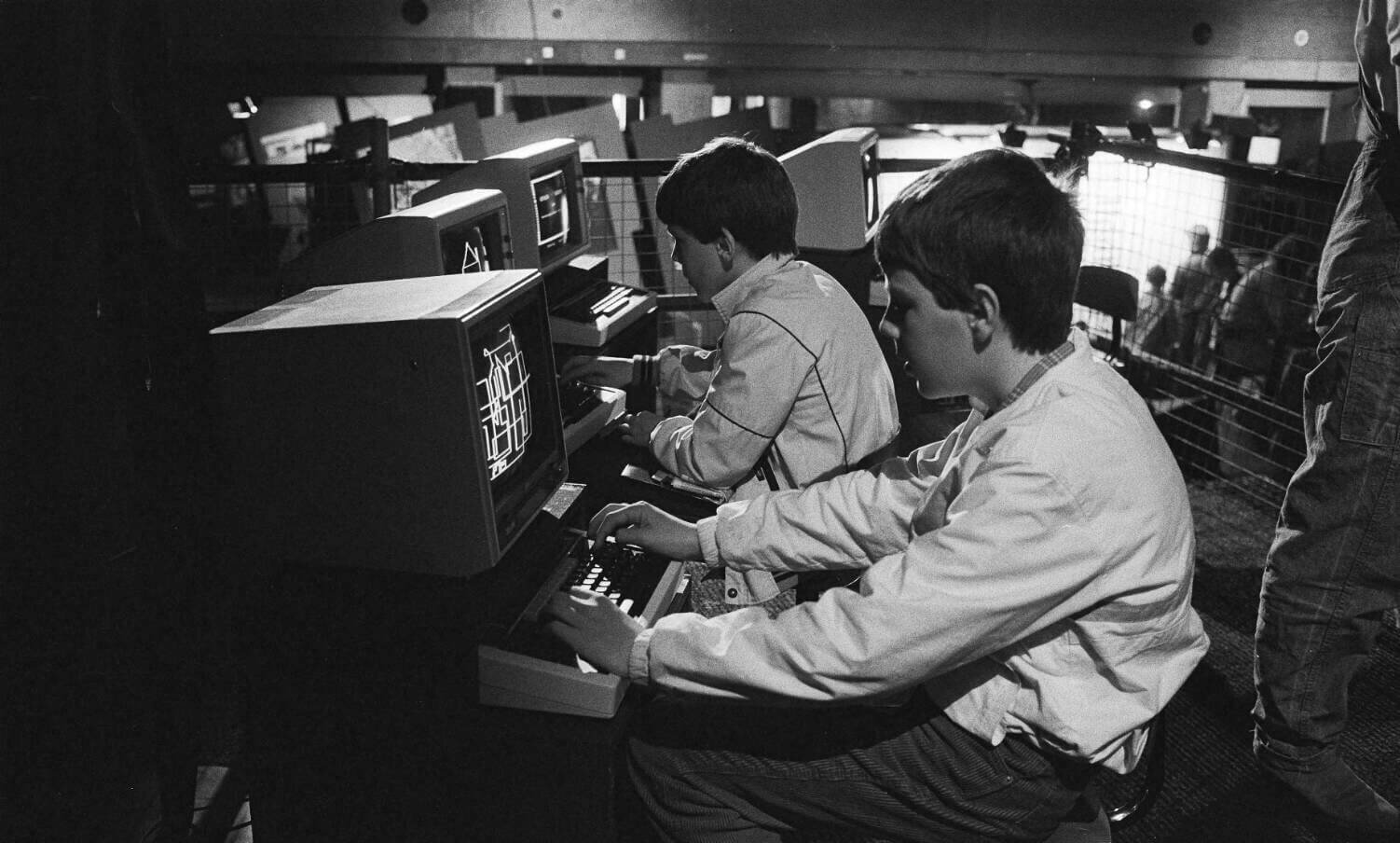 Twee jonge kinderen zitten elk achter een eigen computer op een tafel. Ze kijken allebei naar hun scherm