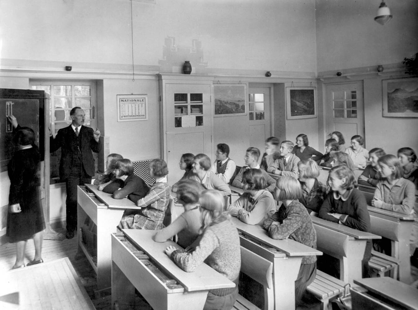 Klaslokaal met schoolklas met meisjes en enkele jongens, circa 1932. Een van de leerlingen tekent op een schoolbord, naast haar de leraar